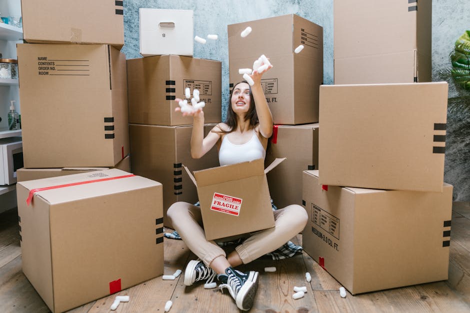 A young woman with long dark hair smiling and holding a large empty cardboard box labeled 'Glass' in red, situated indoors against a textured light blue wall. Several other packed cardboard boxes of v